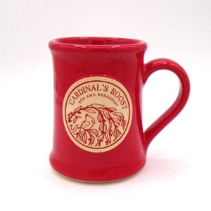 May include: A bright red ceramic mug featuring a circular cream-colored emblem on the front. The emblem displays a line drawing of a bird perched on a branch with the text Cardinal's Roost Bed and Breakfast printed in a circular pattern.