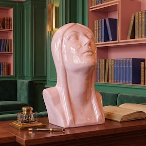 May include: A glossy, light pink ceramic bust sculpture of a person with long, smooth hair and a serene expression, looking upward. The sculpture features a reflective, glazed finish and is positioned on a wooden surface with an inkwell and pen.