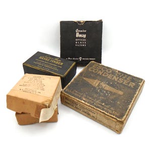 Puede incluir: Una colección de cajas antiguas, incluyendo una con la etiqueta "Genuine Omag Optical Glass Filters" y otra marcada "ESCO Wite-Lite Condenser". Las cajas muestran signos de desgaste, con texto y gráficos visibles. Las cajas son de color negro, marrón y beige.