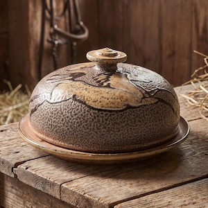 May include: A round ceramic butter dish with a dome-shaped lid featuring a textured, speckled tan and brown finish. The lid has a small, flat circular knob handle at the top, and the base is a matching flat plate with a slightly raised rim edge.