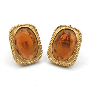 May include: A pair of rectangular gold-toned earrings featuring a central oval-shaped amber-colored faceted stone. The metal frame has a textured rope-style border surrounding the stone, providing a distinct contrast to the smooth, reflective surface of the amber-colored center piece.
