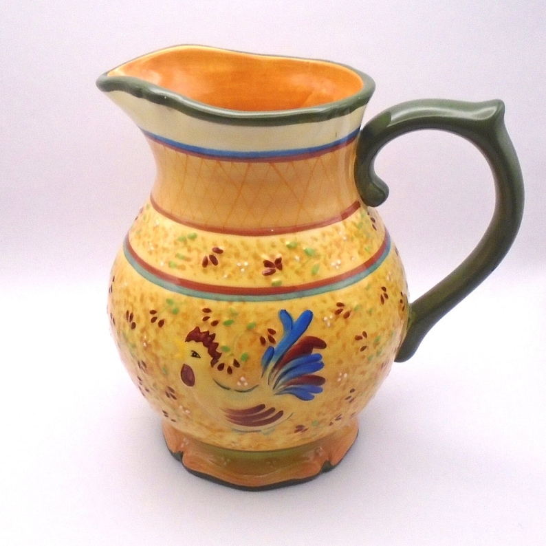 May include: A yellow ceramic pitcher with a green handle and a rooster design. The pitcher has a textured surface and a floral pattern.
