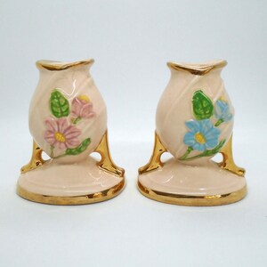 May include: A pair of vintage ceramic bud vases with gold trim and floral designs. The vases are pink with blue and pink flowers and green leaves. The vases have a gold trim around the top and bottom and a gold base.