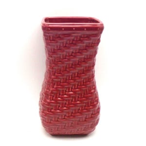 May include: A red ceramic vase with a woven basket design. The vase has a rectangular opening and a slightly curved body. The color is a deep, rich red, and the texture mimics a basket weave.
