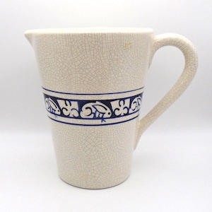 May include: Cream-colored ceramic pitcher with a crackle glaze. A blue band with a repeating floral and bird design encircles the pitcher. The pitcher has a curved handle and a spout for pouring.