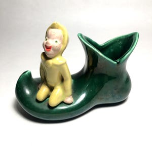 May include: A vintage ceramic figurine of an elf in a yellow outfit with a green-eyed face, seated on a green boot-shaped planter. The planter has a glossy finish, adding to its antique charm. A whimsical decorative piece.