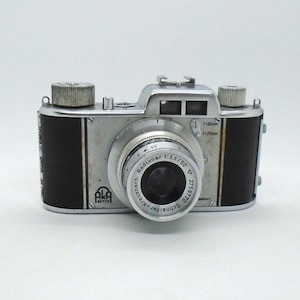 May include: A silver and black 35mm film camera with a black leather covering. The camera has a Schneider Kreuznach Radionar 1:3.5/50 lens and a black lens cap. The camera has a silver body with black accents and a silver lens.