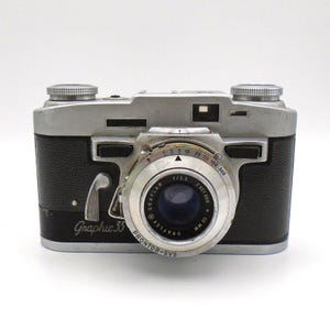 May include: A vintage silver and black Graphic 35 camera. The camera has a silver top and a black leather-like body. The lens has a silver ring with black text and numbers. The camera has two silver dials on top and the text "Graphic 35" on the front.