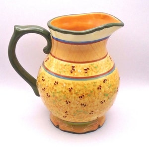 May include: A yellow ceramic pitcher with a green handle and a floral pattern. The pitcher has a wide mouth and a rounded body.
