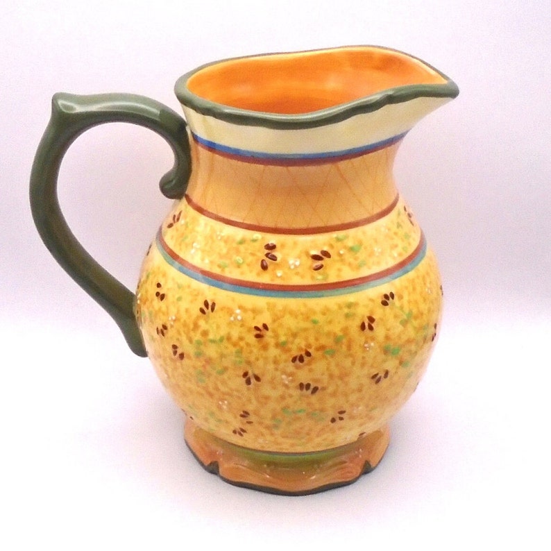 May include: A yellow ceramic pitcher with a green handle and a floral pattern. The pitcher has a wide mouth and a rounded body.