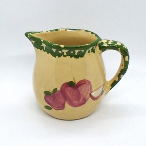 May include: A small, cream-colored ceramic pitcher with a green rim and handle. The pitcher is decorated with hand-painted red apples and green leaves. The pitcher has a spout for easy pouring.