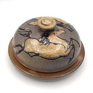 May include: A round ceramic butter dish with a dome-shaped lid featuring a hand-painted horse illustration in shades of tan, brown, and black. The lid has a textured, speckled gray finish and a small circular knob handle on top, resting on a brown base.