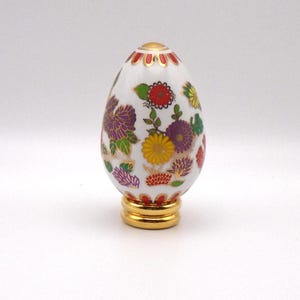 May include: A white porcelain egg with a gold base and a floral design in shades of red, yellow, purple, and green.