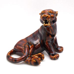 May include: A ceramic tiger figurine in shades of brown, with a glossy finish. The tiger is in a seated position, with its mouth open, revealing its teeth. The figurine has detailed paws and a curled tail.
