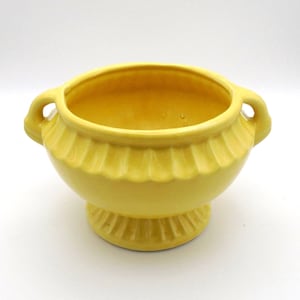 May include: A yellow ceramic bowl with two loop handles and a pedestal base. The bowl has a fluted design around the middle and a smooth interior. The bright yellow color gives the bowl a cheerful and vintage aesthetic.