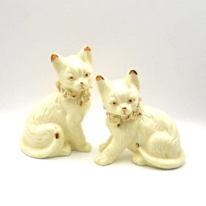 May include: Two cream-colored ceramic cat figurines. Each cat is seated with a floral collar and gold accents on the ears and tail. The cats have a slightly stern expression.