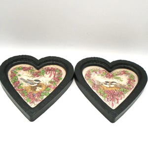 May include: Two heart-shaped framed pictures. Each black frame surrounds a picture of a bird's nest with eggs, surrounded by pink and green floral accents. The pictures are on a white background.