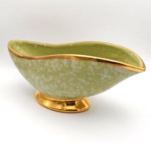 May include: A vintage, light green gravy boat with a speckled pattern and gold trim. The boat has a curved shape and sits on a gold pedestal base. The interior is a solid light green.