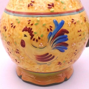May include: Yellow ceramic pitcher with a rooster design. The rooster is painted in shades of red, brown, blue, and yellow. The pitcher has a green and brown trim around the top.