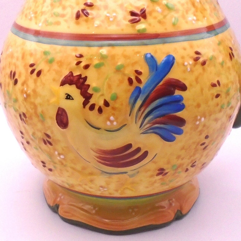 May include: Yellow ceramic pitcher with a rooster design. The rooster is painted in shades of red, brown, blue, and yellow. The pitcher has a green and brown trim around the top.