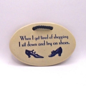 May include: A white oval ceramic sign with blue text that reads "When I get tired of shopping I sit down and try on shoes."  Two blue shoes are pictured on the sign.