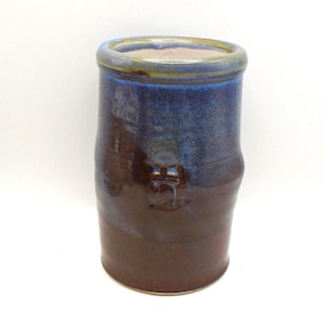 May include: A handmade ceramic vase with a cylindrical form. The vase displays a gradient of colours, shifting from a deep brown at the base to a vibrant blue at the top. A small, textured detail is present on the side.