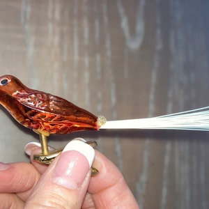 Puede incluir: Un pequeño adorno de pájaro de vidrio con un pico rojo, un cuerpo marrón y plumas de cola blancas. El pájaro está sentado en un soporte dorado.