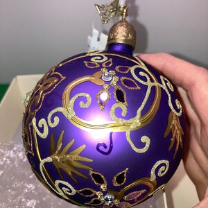 May include: Purple glass Christmas ornament with gold glitter and a gold and white floral design. The ornament has a gold top with a butterfly design.