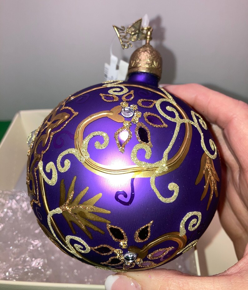 May include: Purple glass Christmas ornament with gold glitter and a gold and white floral design. The ornament has a gold top with a butterfly design.