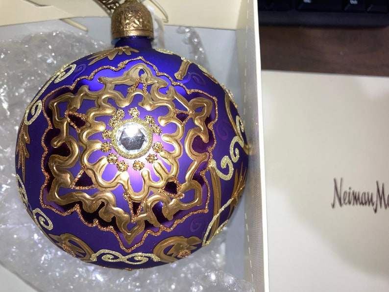May include: A purple glass Christmas ornament with gold detailing and a clear rhinestone in the center. The ornament is in a white box with the text "Neiman Marcus" printed on the bottom.