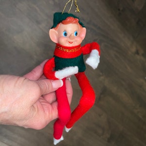 Könnte beinhalten: Ein Weihnachts-Elfen-Anhänger aus Filz mit grünem Hut, roter Hose und weißen Handschuhen. Der Elf hat ein lächelndes Gesicht mit blauen Augen und roten Haaren.