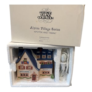 May include: A Heritage Village Collection Alpine Village Series porcelain building titled Apotek and Tabak, featuring a blue roof, cream walls, and brown shutters. The set includes a white light cord and is housed in its original cardboard box.