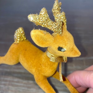 Pode incluir: Uma figura de rena de veludo marrom com chifres de glitter dourado e uma fita dourada em volta do pesco&ccedil;o.
