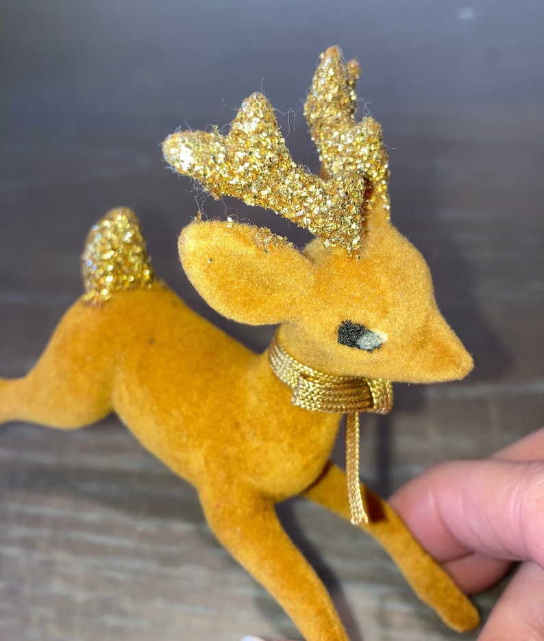 Pode incluir: Uma figura de rena de veludo marrom com chifres de glitter dourado e uma fita dourada em volta do pesco&ccedil;o.