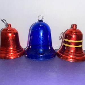Op de afbeelding: Drie kerstbellen, twee rode en één blauwe. De rode bellen hebben een metallic afwerking, en één heeft gele strepen. De blauwe bel heeft een glad, glanzend oppervlak. Een feestelijke decoratie.