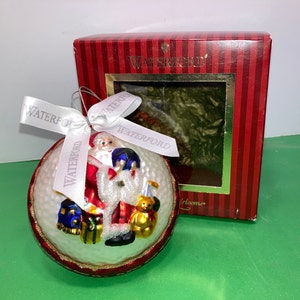 Puede incluir: Adorno navideño de vidrio blanco con ribete rojo y dorado. El adorno presenta una figura de Papá Noel sosteniendo un juguete y un oso de peluche. El adorno está en una caja de rayas rojas y blancas con la palabra "Waterford" impresa en ella.