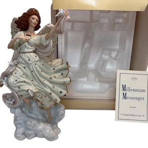 Op de afbeelding: Een porseleinen engelenbeeldje, de "Millennium Messenger" van Lenox, in een witte jurk met gouden sterren. De engel houdt een duif vast en rust op een wolkenbasis. Het beeldje zit in de originele verpakking. Het label luidt "Limited Edition Issue".