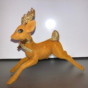 Pode incluir: Uma figura de rena de veludo marrom com chifres de glitter dourado, uma cauda de glitter dourado e uma fita dourada em volta do pesco&ccedil;o.