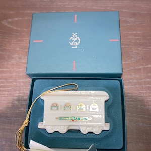 May include: A white porcelain Christmas ornament shaped like a vintage trolley car with gold accents and green trim. The ornament has the text "1991 Lenox" on the side. The ornament is in its original box with a blue lid that says "Lenox" in gold lettering.