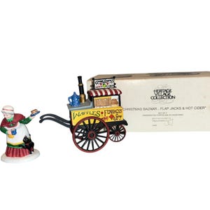 May include: A Heritage Village Collection Christmas Bazaar set featuring a miniature porcelain figure of a woman and a food cart. The cart is yellow with red and black accents, and the woman is wearing a red and green outfit.