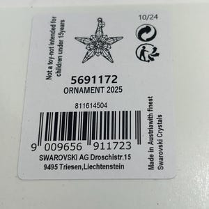 May include: A white product tag with a star-shaped ornament design. The tag includes the text "ORNAMENT 2025", a barcode, and the text "Made in Austria with finest Swarovski Crystals".