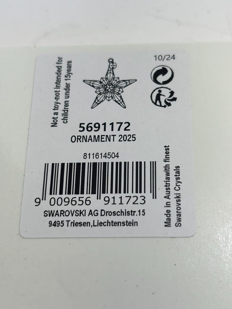 May include: A white product tag with a star-shaped ornament design. The tag includes the text "ORNAMENT 2025", a barcode, and the text "Made in Austria with finest Swarovski Crystals".