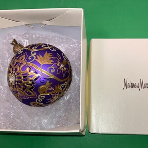 May include: A purple and gold Christmas ornament with a floral design and rhinestones in a white box with the text "Neiman Marcus" on the lid.