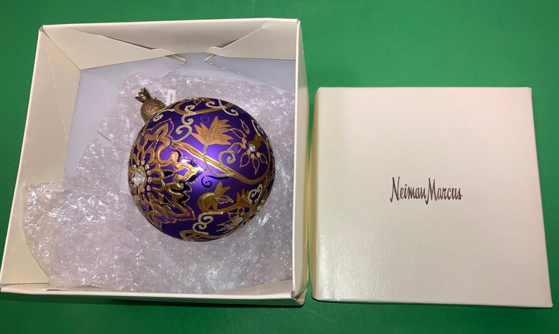 May include: A purple and gold Christmas ornament with a floral design and rhinestones in a white box with the text "Neiman Marcus" on the lid.