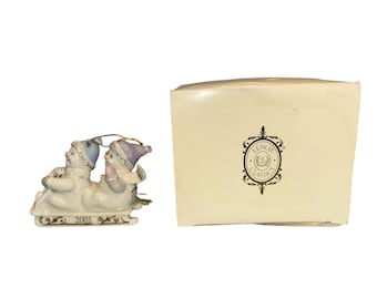 May include: A Lenox Classics ornament depicting two figures in winter hats and scarves on a sled. The ornament is white with blue and purple accents. The year 2001 is inscribed on the sled. A cream-coloured box with the Lenox Classics logo is next to the ornament.