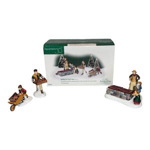 May include: Department 56 Tending The Cold Frame figurine set including a stone cold frame, three figures, and a wheelbarrow. The packaging box features the product name and a graphic illustration of the set in a snowy garden scene.