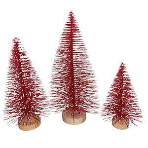 Può includere: Tre alberi di Natale rossi a spazzola di diverse dimensioni. Ogni albero ha una base in legno ed è ricoperto di setole rosse con una spolverata di neve bianca. Gli alberi sono disposti su uno sfondo bianco.