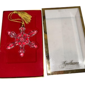 Puede incluir: Adorno navideño de copo de nieve rojo con borla dorada, presentado en una caja forrada de terciopelo rojo con ribete dorado. La caja está abierta, revelando el adorno y el logotipo de Gorham.