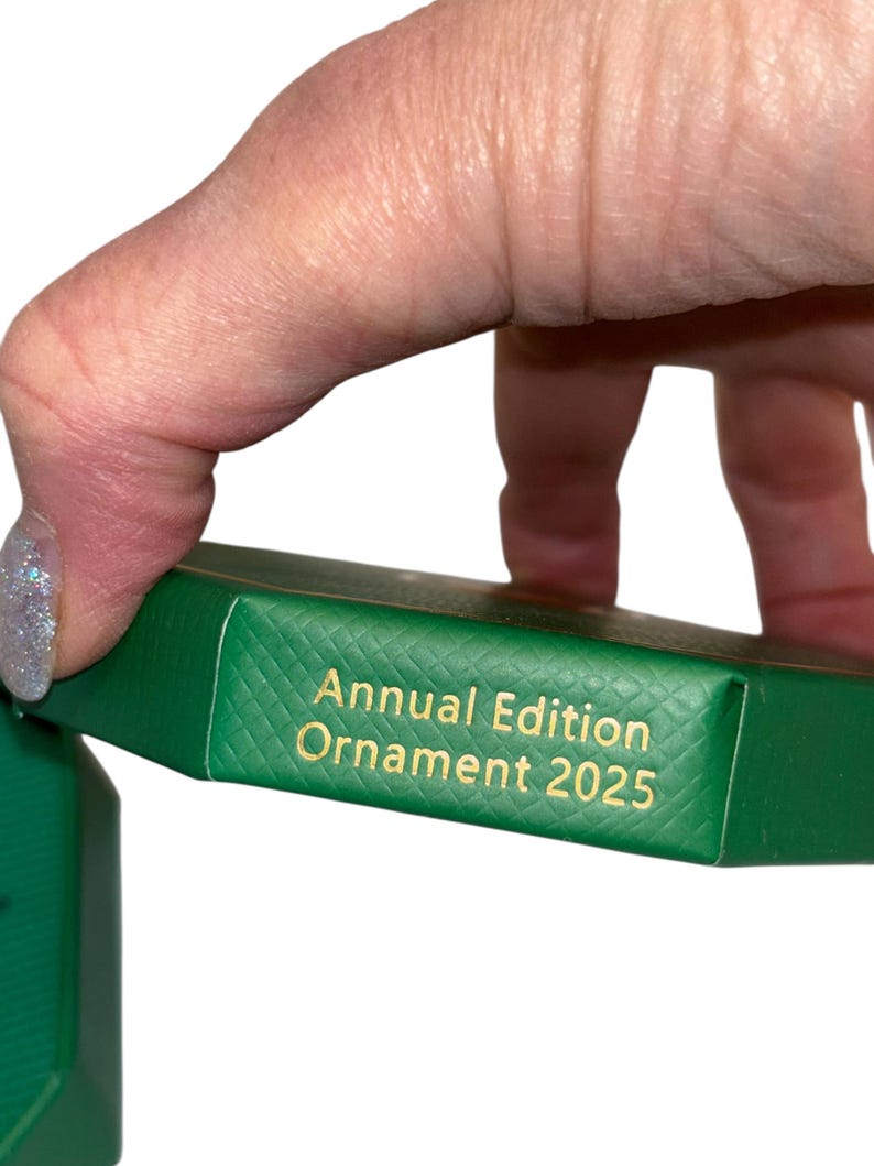 May include: A green box with gold text that reads "Annual Edition Ornament 2025." The box is held by a hand with a glittery nail. The box has a textured surface.