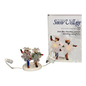 May include: A Department 56 Snow Village lit accessory titled Brite Lites Christmas Parade Marching Snowflakes. The set features two figures wearing large white snowflake costumes with glowing centers, attached to a white base with a power cord.